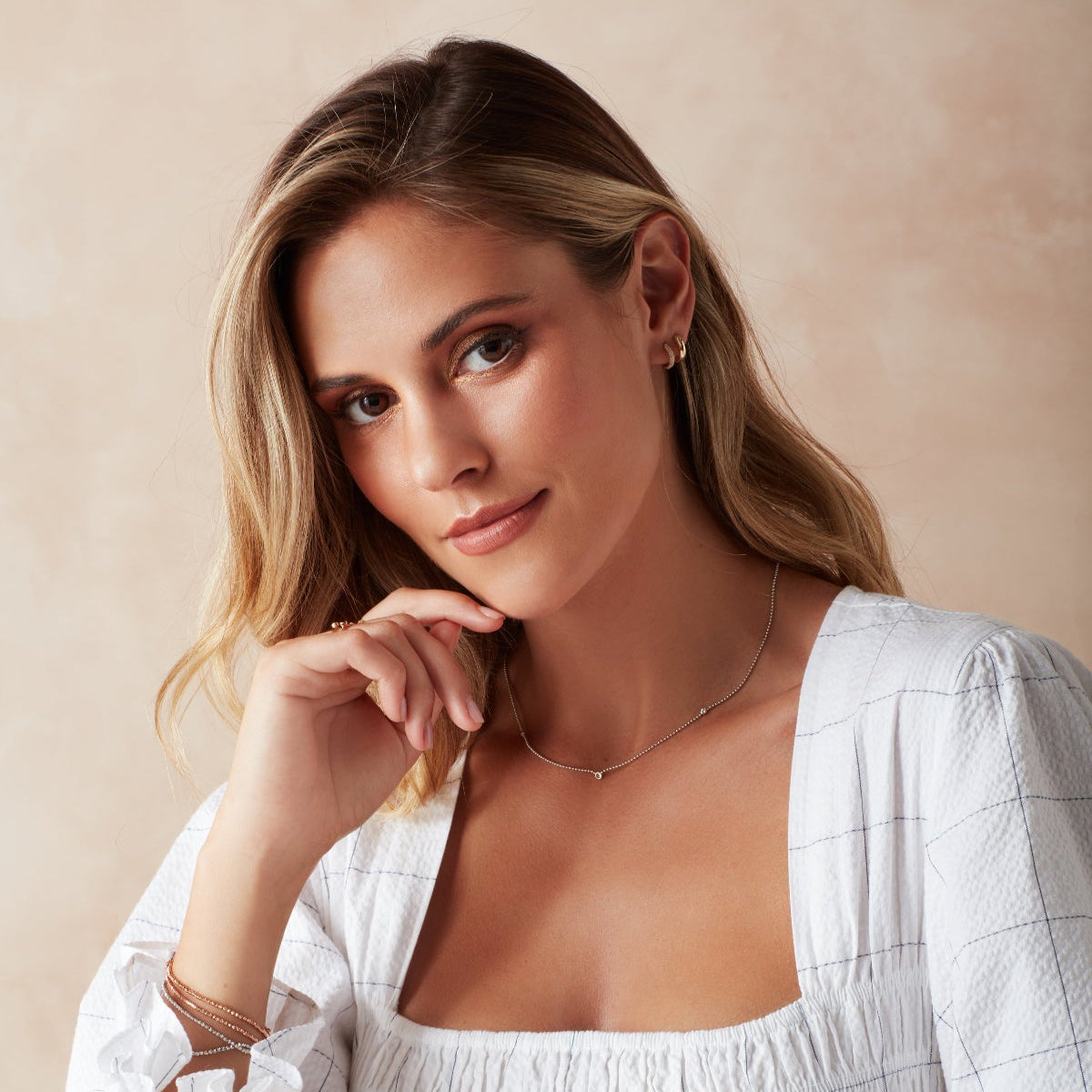 A blonde woman wearing a white gold Triple Diamond Layering Necklace, a Gold Huggie, a Pavé Huggie, and several rings and bracelets, including Diamond Cut Bracelets in white and rose gold, The Slide Chain, a Tres Pearl Ring and Delicate Trio Stacking Rings, styled with a white dress against a light neutral background.