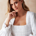 A blonde woman wearing a white gold Triple Diamond Layering Necklace, a Gold Huggie, a Pavé Huggie, and several rings and bracelets, including Diamond Cut Bracelets in white and rose gold, The Slide Chain, a Tres Pearl Ring and Delicate Trio Stacking Rings, styled with a white dress against a light neutral background.