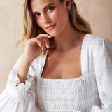 A blonde woman wearing a white gold Triple Diamond Layering Necklace, a Gold Huggie, a Pavé Huggie, and several rings and bracelets, including Diamond Cut Bracelets in white and rose gold, The Slide Chain, a Tres Pearl Ring and Delicate Trio Stacking Rings, styled with a white dress against a light neutral background.