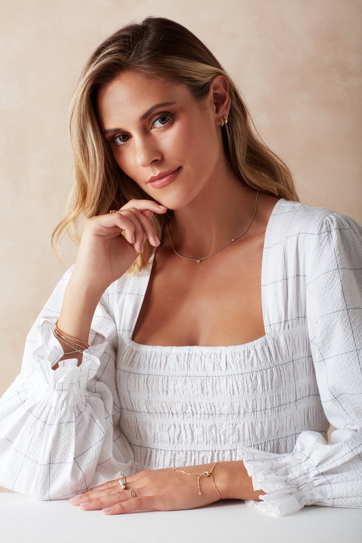 A blonde woman wearing a white gold Triple Diamond Layering Necklace, a Gold Huggie, a Pavé Huggie, and several rings and bracelets, including Diamond Cut Bracelets in white and rose gold, The Slide Chain, a Tres Pearl Ring and Delicate Trio Stacking Rings, styled with a white dress against a light neutral background.