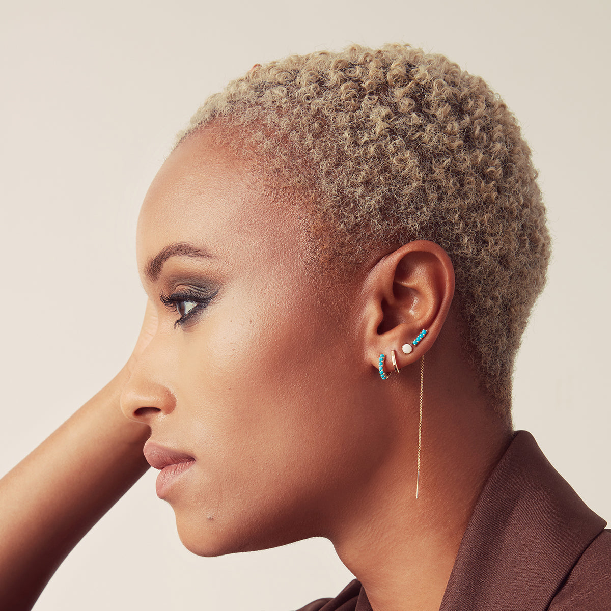 An Alli Webb Turquoise Huggie, Faceted Huggie, Opal Comfort Stud and Turquoise Threader worn by a woman with short blonde hair and a brown top against a light background. 