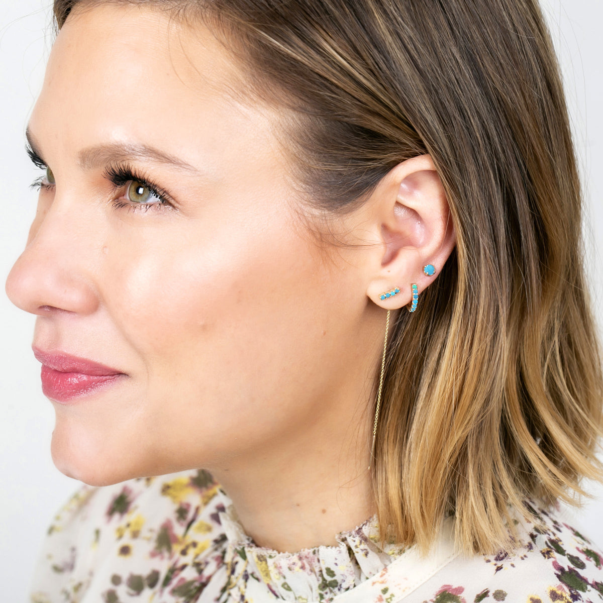 Close-up of a woman wearing an Alli Webb Turquoise Threader, Turquoise Huggie and Comfort Stud, with blonde short hair and a printed top against a light grey background. 