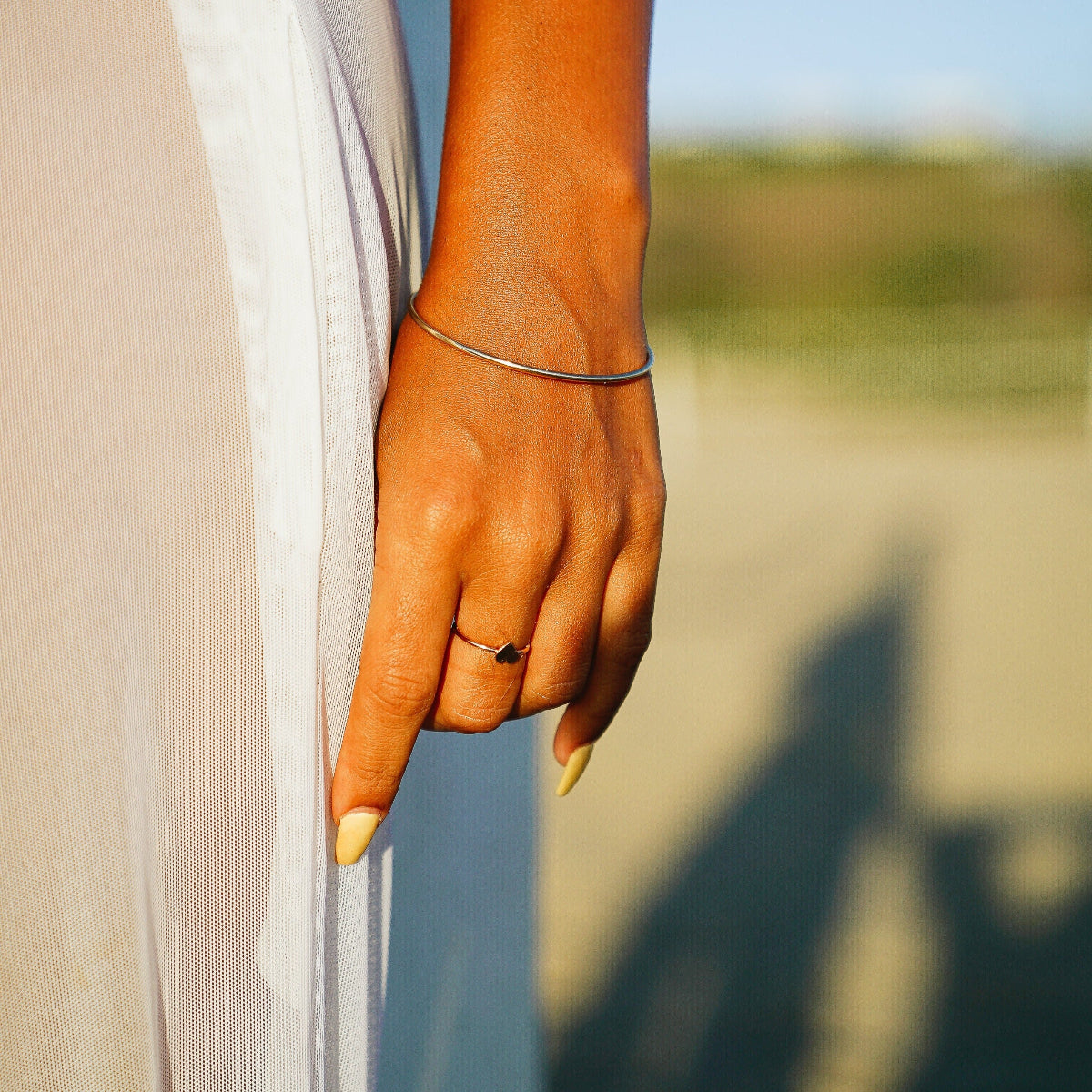 A close-up of a woman’s hand wearing a white gold Simple Bangle and a Heartfelt Ring, resting on a white sheer dress against a blurred background.