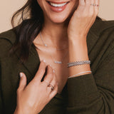 A woman wearing two Rolo Bracelets and two Diamond Cut Bracelets, a 14k Gold Asymmetrical Pavé Heart Charm Necklace and a Pavé MAMA Necklace, all in white gold, and several rose gold rings, in a dark khaki green knit top.