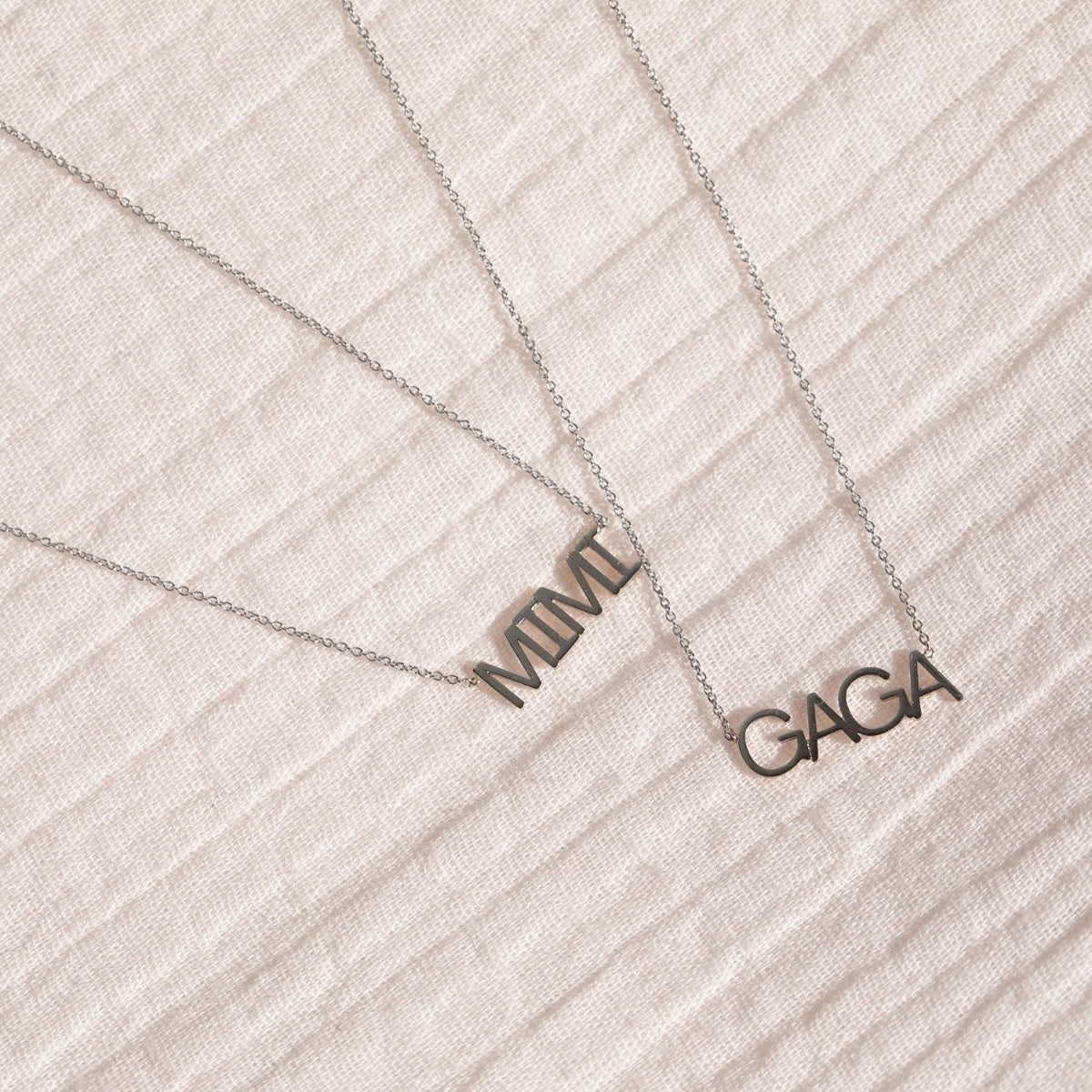 A white gold MIMI Necklace and GAGA Necklace against an off-white fabric background. 