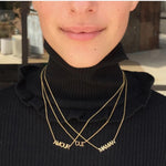 A close-up of a person wearing three yellow gold word plate necklaces in AMOUR, OUI and MAMAN, and a black turtle neck top.