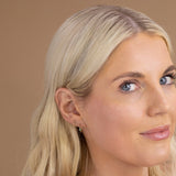 A blonde woman wearing a yellow gold Birthstone Hoop in Emerald - May and Open Eclipse Earring - Mini against a light brown background. 