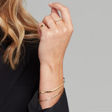 A woman's hand wearing an Overboard Bracelet, another gold bracelet, a Braided Ring, a Delicate Beaded Band and a Perla Pinky Ring, styled with a black blazer. 