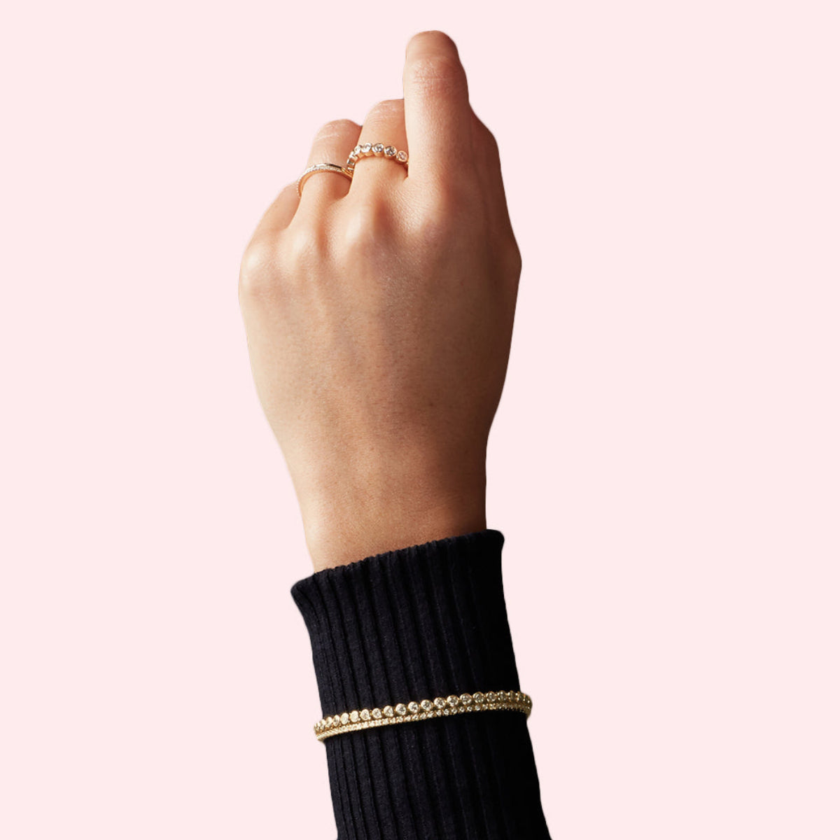 A Love Set Match Bracelet, a Down the Line Bracelet and two rings, including The Evermore Ring, worn on a woman’s arm held up in a black long sleeve against a pale pink background.