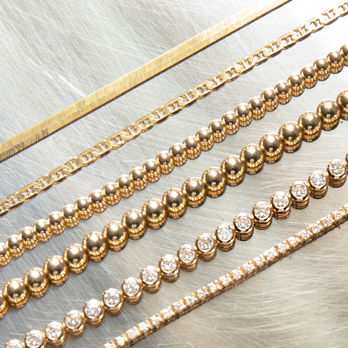 Multiple yellow gold bracelets laid against  a silver metal surface, being an Overboard Bracelet, Bubble Bracelets (Large and Small), a Love Set Match Bracelet and a Down the Line Bracelet. 