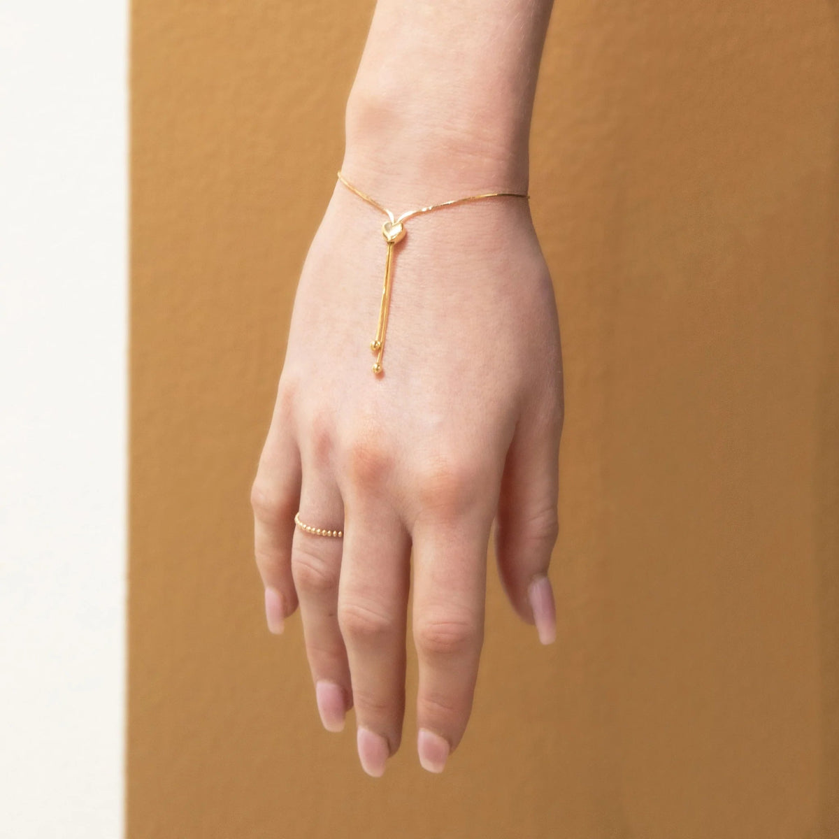 A hand facing down wearing The Slide Heart Bracelet and Delicate Beaded Band in yellow against a tan brown background. 