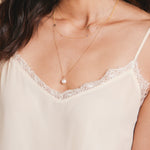A close-up of a décolletage wearing a 14k Gold Asymmetrical Charm Necklace - Angel Wing, a Custom Gold Necklace - 4 Letters and a Perla Necklace, all in yellow gold, styled in a cream lace strap top. 