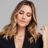 A blonde woman wearing an Overboard Layering Chain, a Sequence Chain, an Element Long Link Chain, a Perla Pinky Ring and a Charmed I'm Sure Hoop, all in yellow gold, with a black blazer against a light grey background.