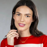 A woman wearing a MAMAN Necklace, a Straight as an Arrow Earring, a Link Ring, a Delicate Band, a Braided Ring and Geo Stacking Rings, all in yellow gold, with brown hair and a red and white jumper against a light grey background.