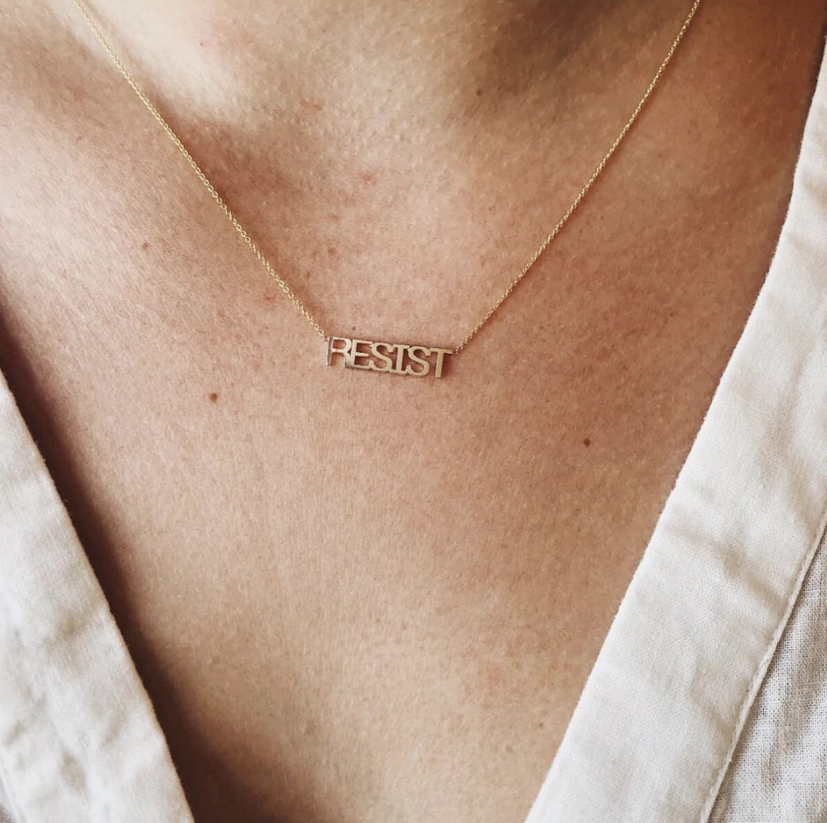 A close-up of a décolletage wearing a RESIST Necklace with a cream linen top. 