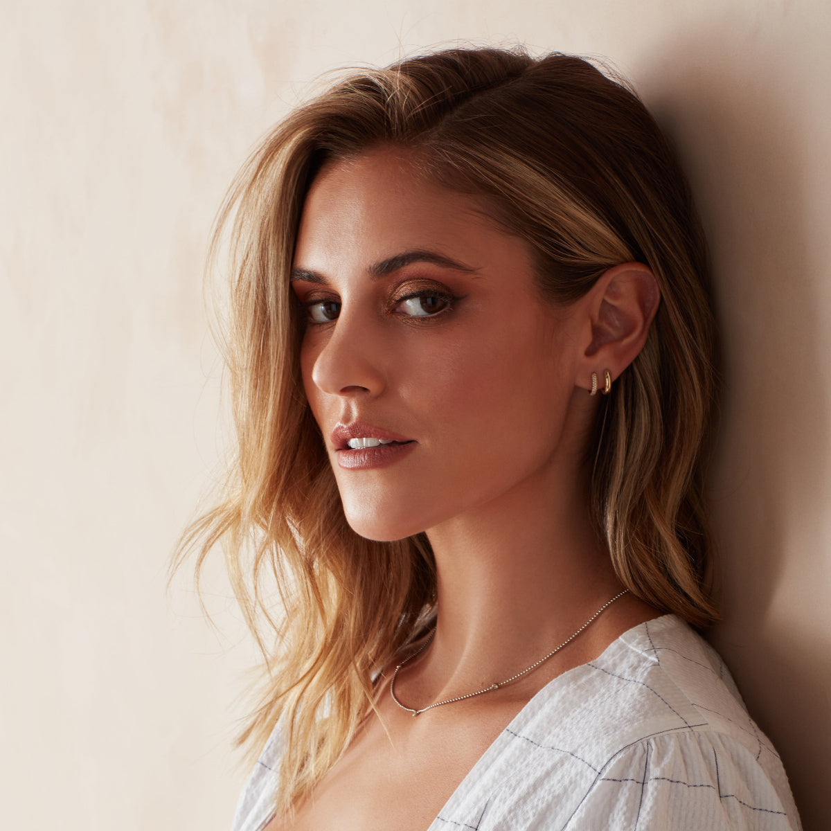 A blonde woman wearing a Gold Huggie, Pave Huggie and Triple Birthstone Layering Necklace - White Gold, with a white top against a light beige background. 