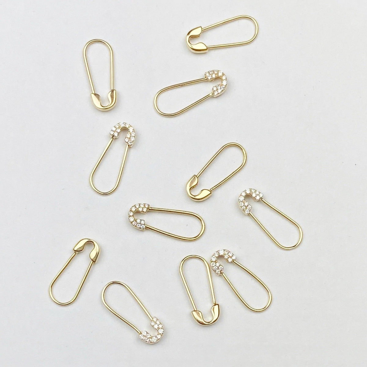 Several Gold Safety Pin Earrings and Pavé Safety Pin Earrings against a light grey background. 