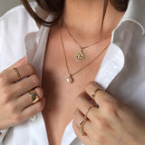 Close-up of a brunette woman's décolletage wearing a Zodiac Necklace - Leo and a Perla Necklace, with her hands wearing a Link Ring, a Thick Band, Birthstone Rings, and a Geo Stacking Ring Square, in a white shirt.