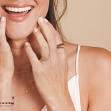 A close-up of a woman smiling with her hands on her face, wearing an Etched Paris Ring, Delicate Trio Stacking Rings, yellow gold necklaces and two Bouquet bracelets, styled in a white top.
