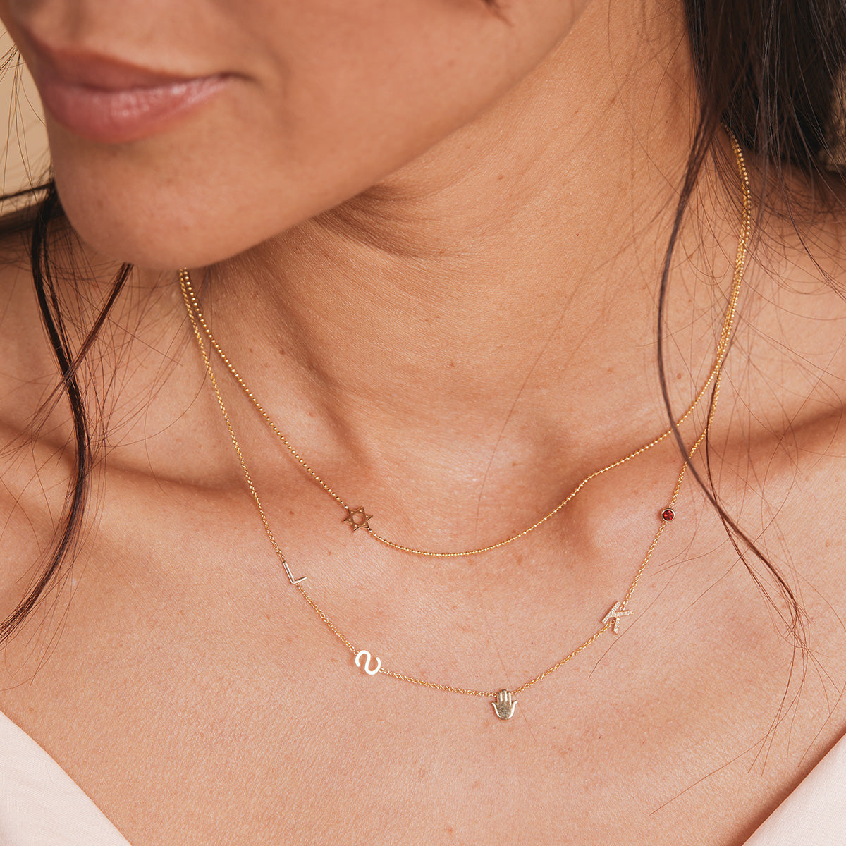 A close-up of a woman’s neck wearing a 14k Gold Asymmetrical Charm Necklace - Star of David with a bead chain, and a Custom Gold Necklace - 5 Letters (L, S, a hand, K and a birthstone), both in yellow gold.